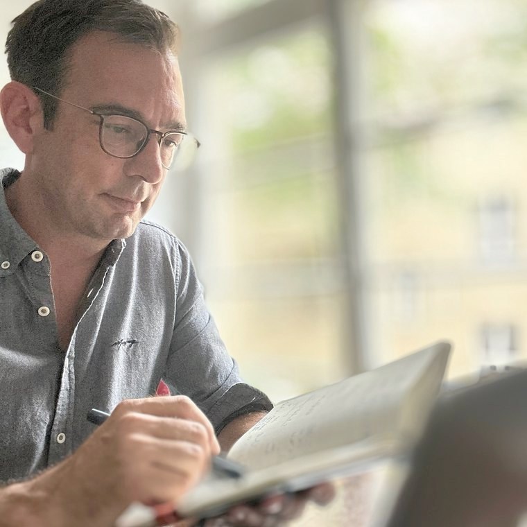 Thomas Kohns im grauen Hemd schreibt am Fenster in ein Notizbuch