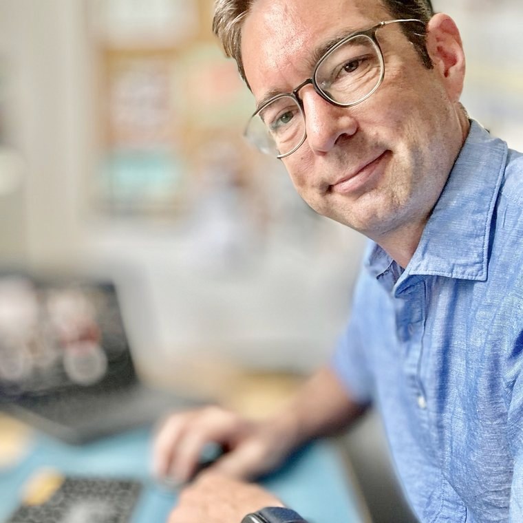 Thomas Kohns sitzt am Schreibtisch vor dem Laptop, die Hand an der Maus, und schaut in die Kamera